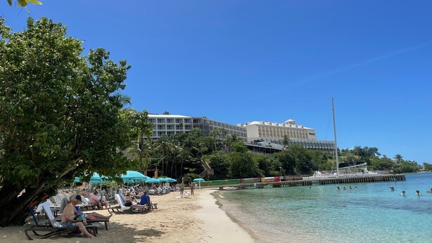 Ein Strand mit Liegestühlen und Sonnenschirmen, ein Hotel im Hintergrund und ein Steg, der sich ins Wasser erstreckt.