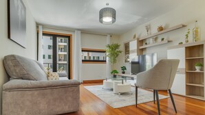 Un salon moderne avec un canapé, un fauteuil, une table basse en verre et un téléviseur. Le sol est en bois, il y a un luminaire au plafond et des étagères avec des plantes.