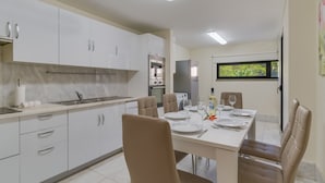 Une cuisine moderne avec des meubles blancs, un coin repas avec une table dressée pour quatre personnes et une fenêtre donnant sur un paysage verdoyant.