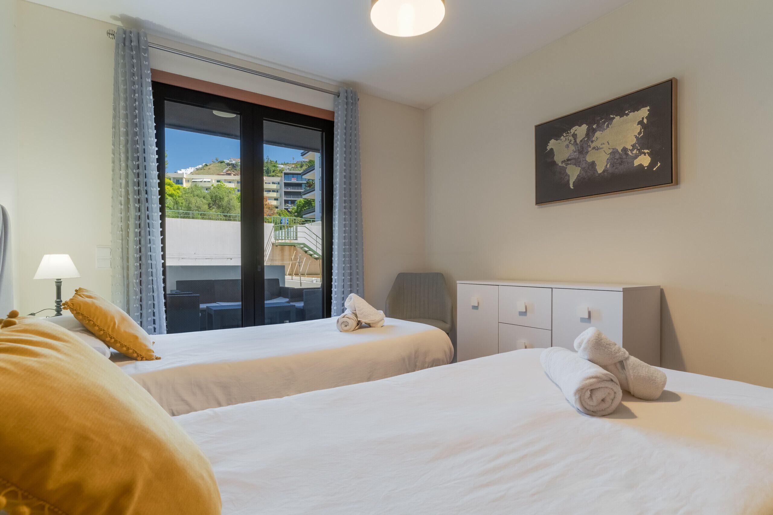 Ein Hotelzimmer mit zwei Betten, einem Schreibtisch, einem Sessel, einer Lampe, einer Karte an der Wand und Blick ins Freie.