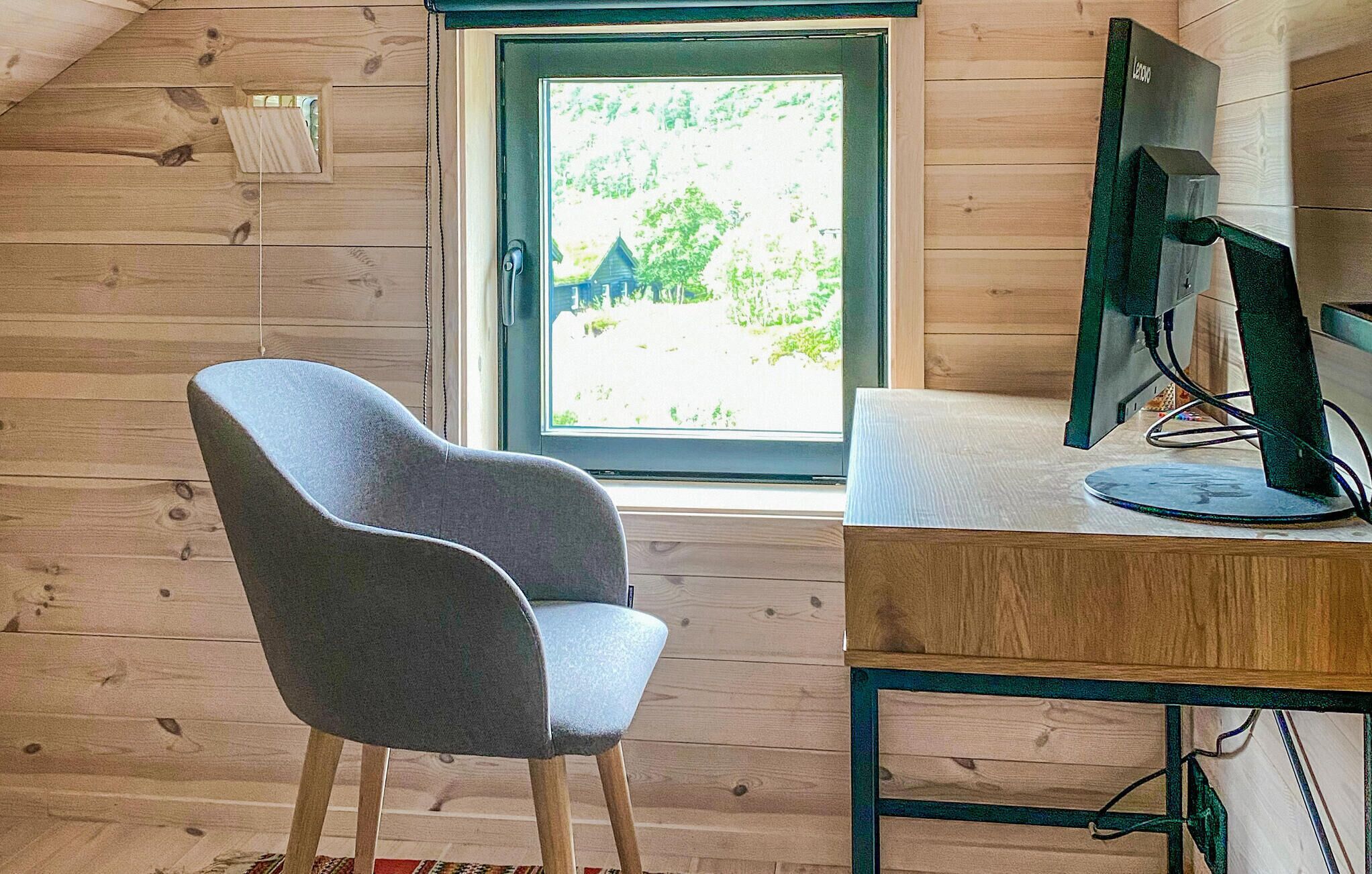 Ein Zimmer mit Holzbalkendecke, einem Fenster mit Blick auf Grünflächen, einem grau bezogenen Sessel und einem Schreibtisch mit Computermonitor.