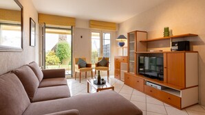 Un salon moderne comprenant un canapé, une table basse, un téléviseur et un meuble. Une porte donne accès à un balcon d’où l’on peut admirer la verdure.