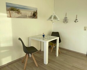 A small dining area with a white table and two chairs, a painting of a beach scene, a hanging lamp, and a wall-mounted clock. - Studioapartment in der Frische Brise 04.05 - Frische Brise (Cuxhaven)
