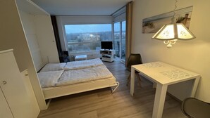 A hotel room with a bed, a desk, a chair, a TV, and a large window offering a city view. - Studioapartment in der Frische Brise 04.05 - Frische Brise (Cuxhaven)
