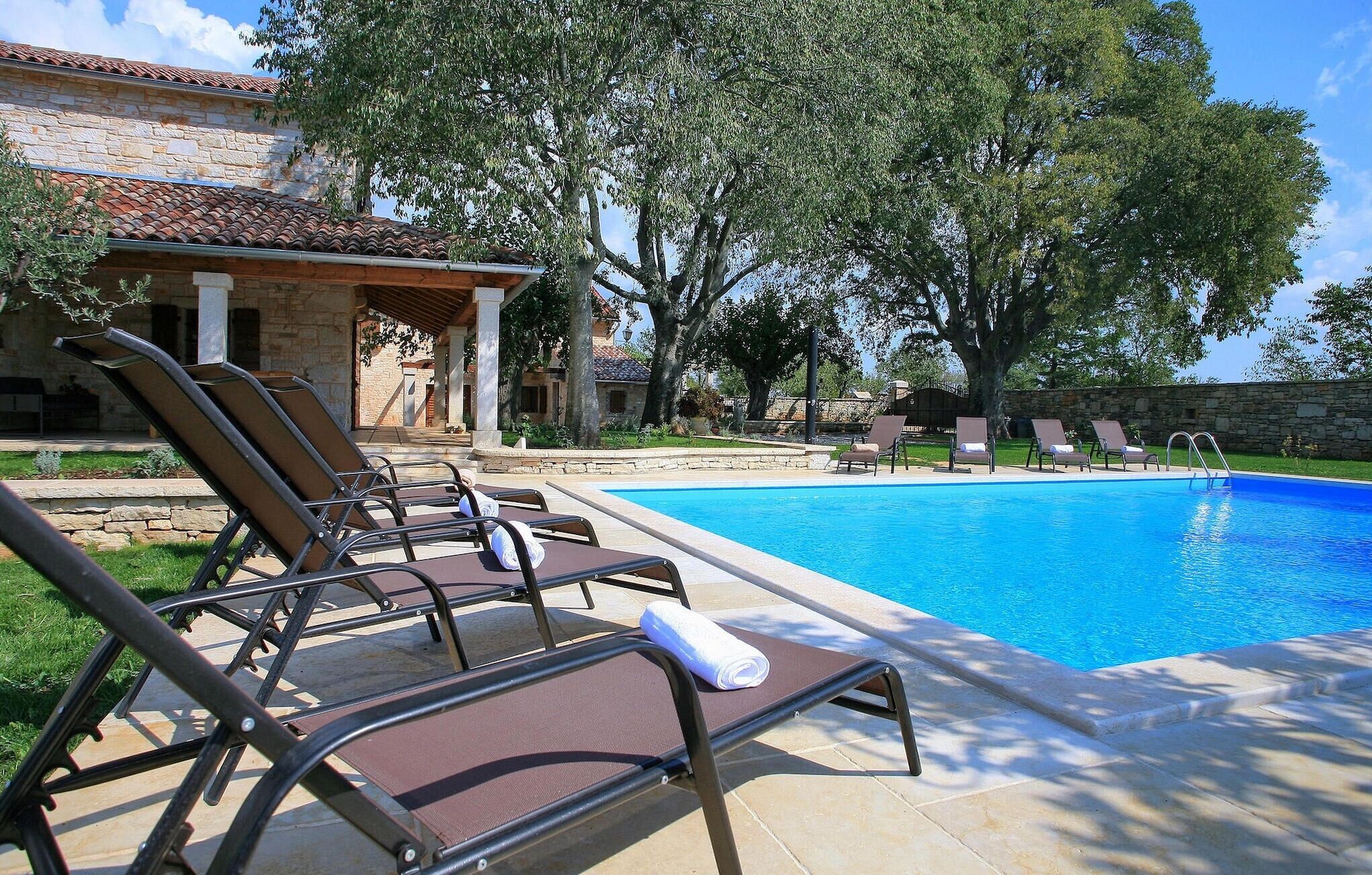 Zona de piscina con sillones reclinables y un edificio de piedra al fondo.