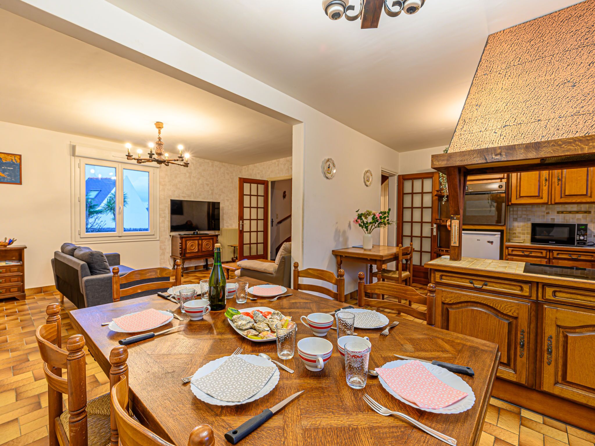 Un coin repas avec une table en bois dressée pour quatre personnes, entourée de chaises en bois. Un téléviseur est fixé au mur, et on aperçoit, au fond, une cuisine avec des meubles en bois.