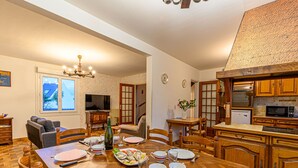 Zona pranzo con tavolo in legno apparecchiato per quattro, circondato da sedie in legno. Una televisione è montata a parete, e sullo sfondo si intravede una cucina con mobili in legno.