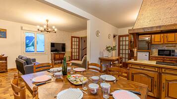 Un coin repas avec une table en bois dressée pour quatre personnes, entourée de chaises en bois. Un téléviseur est fixé au mur, et on aperçoit, au fond, une cuisine avec des meubles en bois.
