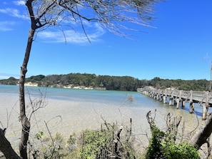 A clear view of a beach with a wooden pier extending into the water, surrounded by trees and clear skies. - The Bungalow - Luxurious & Calm | Beach & Bush (Valla Beach)