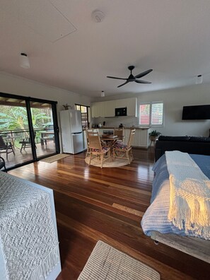 A modern kitchen with white cabinets, a dining area with a round table and chairs, a ceiling fan, and a large sliding glass door leading to an outdoor area. - The Bungalow - Luxurious & Calm | Beach & Bush (Valla Beach)