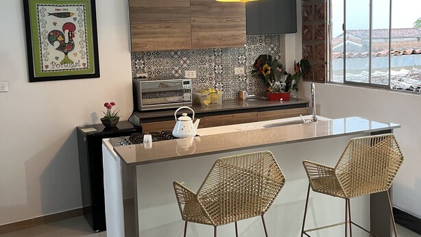 A modern kitchen with a central island, two wicker bar stools, a red pendant light, and a large window with a view.