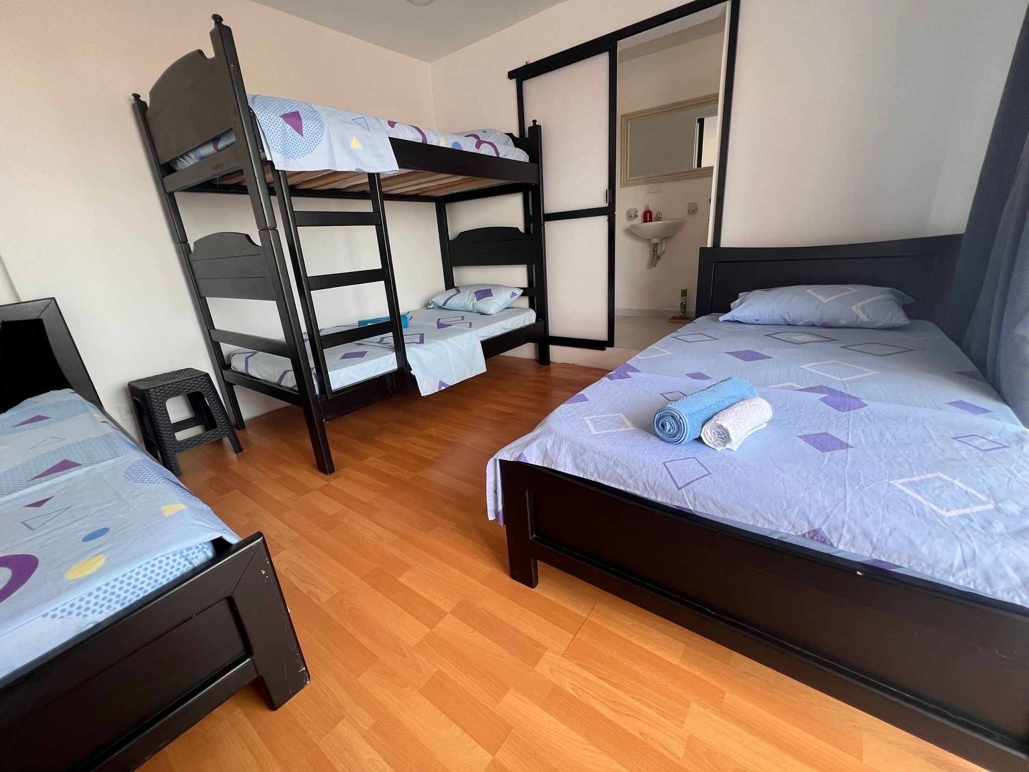A room with two beds, one with a headboard and one with a bunk bed. There is a small stool and a mirror on the wall.