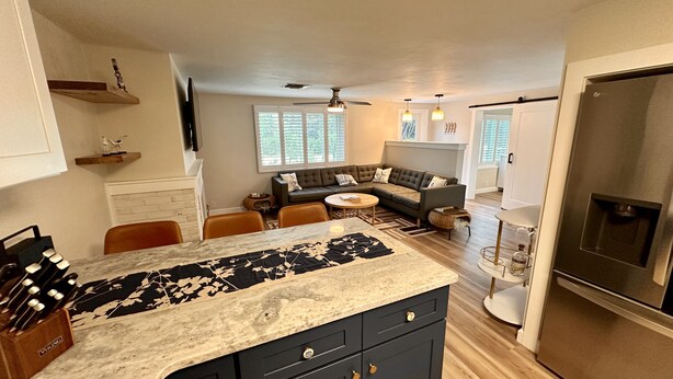 A modern kitchen with dark cabinetry, a marble countertop, and a built-in oven. There is a dining area with a round table and chairs, and a living room with a sectional sofa and a fireplace.