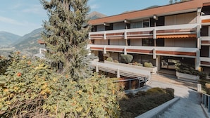 A multi-story building with balconies, surrounded by greenery and mountains in the background.
