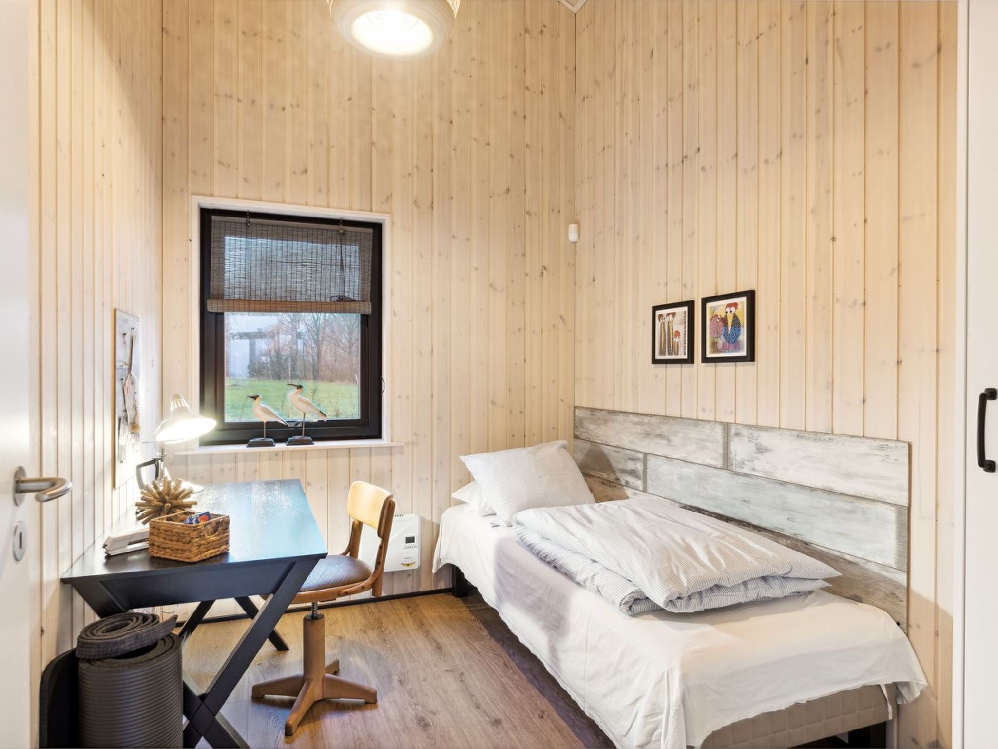 Una habitación con cama, un escritorio con silla, una ventana con vistas al exterior y dos cuadros en la pared.