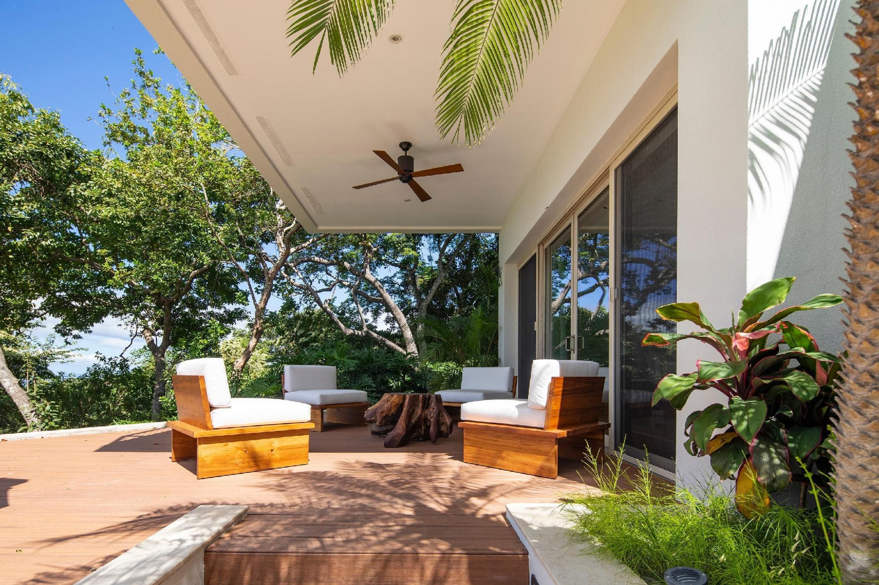 A modern outdoor patio with wooden furniture, a ceiling fan, and lush greenery.