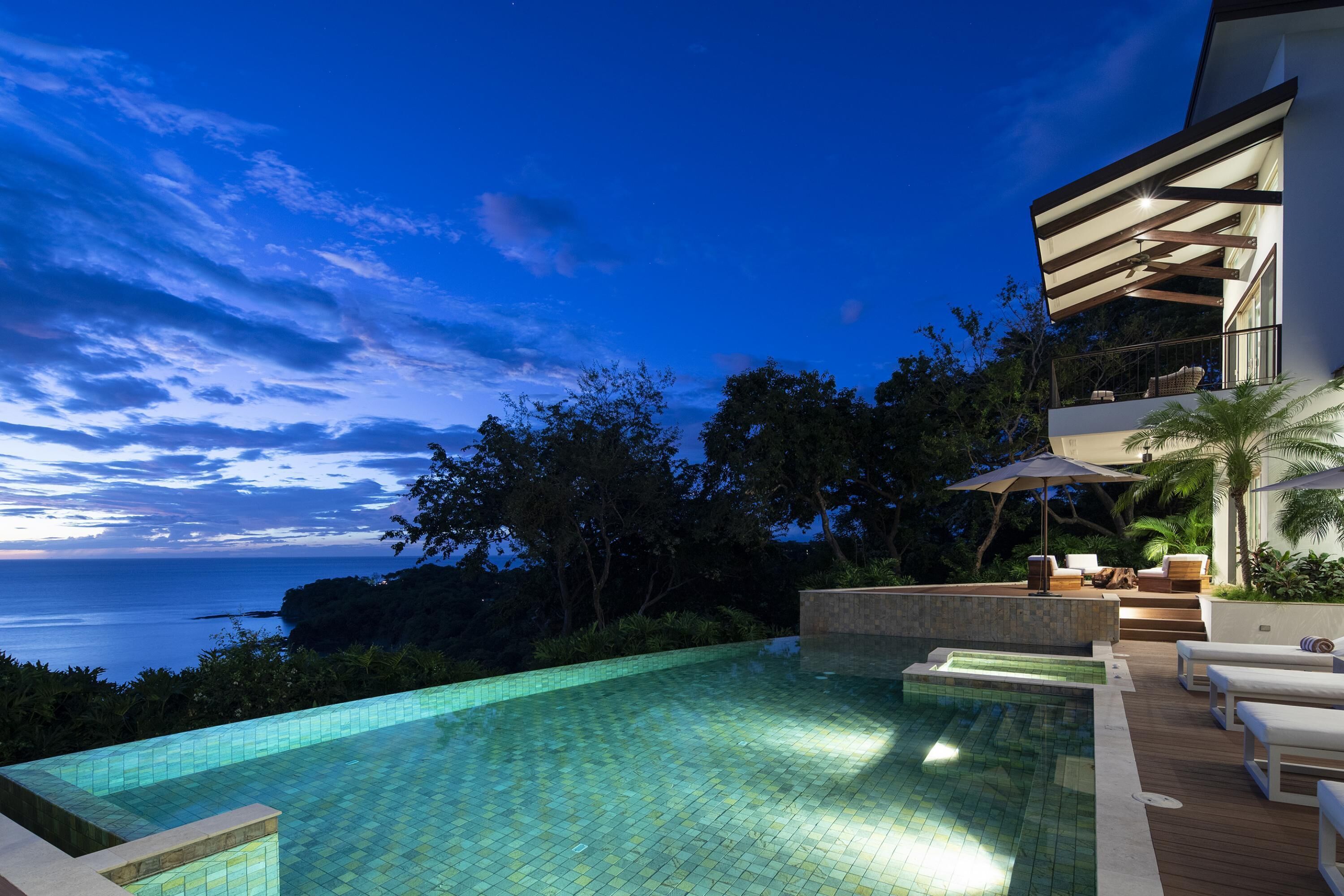 A swimming pool with a view of the ocean, surrounded by lush greenery and modern outdoor furniture.