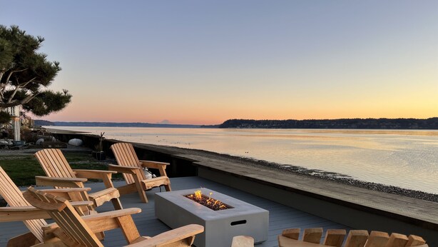 Terraza con sillas Adirondack de madera y un brasero con vista a un cuerpo de agua tranquilo al atardecer.