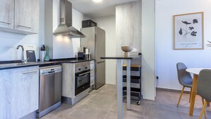 A modern kitchen with stainless steel appliances, a built-in oven, and a dishwasher. There is a small table with a bowl on it and a chair.