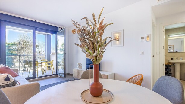 A modern living room with a round table, a vase with dried flowers, a large window with a view of the outdoors, and a dining area with yellow chairs.