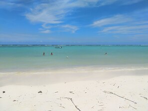 Une plage à l’eau turquoise et limpide, un bateau à l’horizon et quelques personnes qui se baignent.