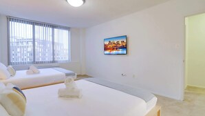 Ein Hotelzimmer mit zwei Betten, einem großen Fenster mit Jalousien, einem Stadtbild an der Wand und einer Tür rechts.