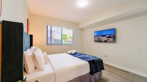 A hotel room with a bed, a window with blinds, a cityscape painting, and a ceiling light.