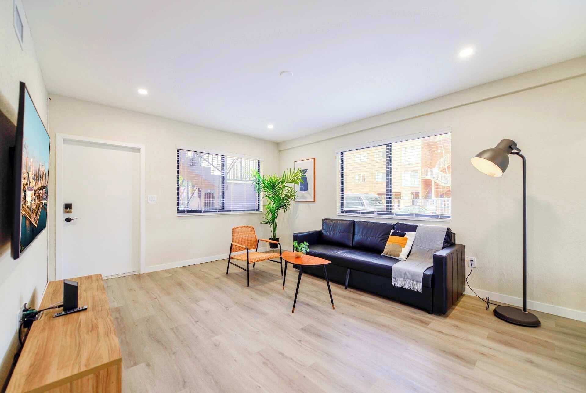 Un salon moderne avec un canapé en cuir noir, une table basse en bois et un petit fauteuil orange. Une grande fenêtre avec des stores, une lampe sur pied et un tableau encadré ornent le mur.