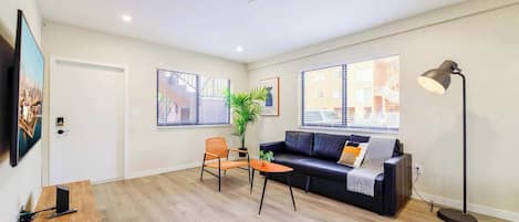 Un salon moderne avec un canapé en cuir noir, une table basse en bois et un petit fauteuil orange. Une grande fenêtre avec des stores, une lampe sur pied et un tableau encadré ornent le mur.