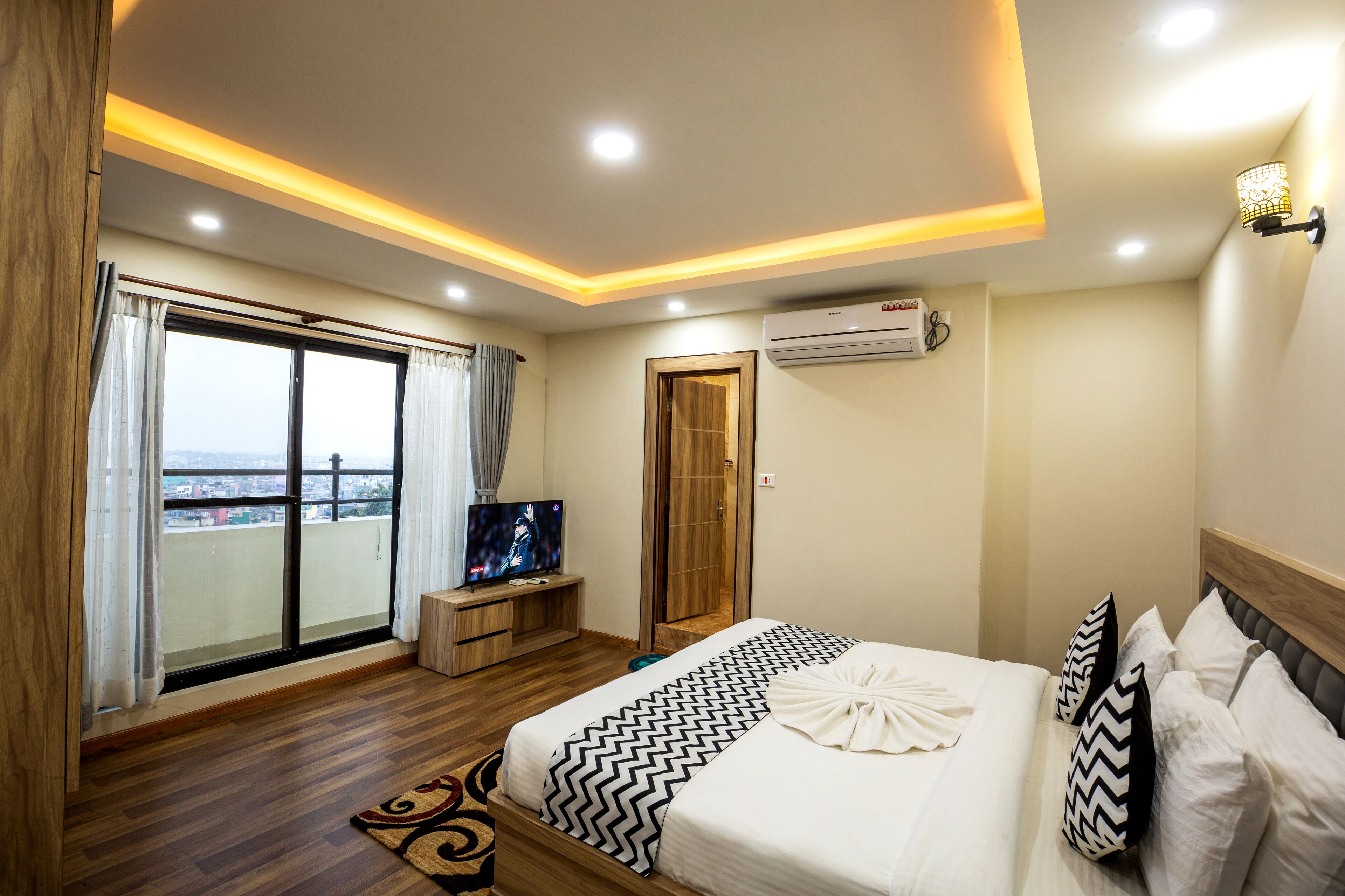 A hotel room with a large bed, a TV, a balcony with curtains, and an air conditioning unit.