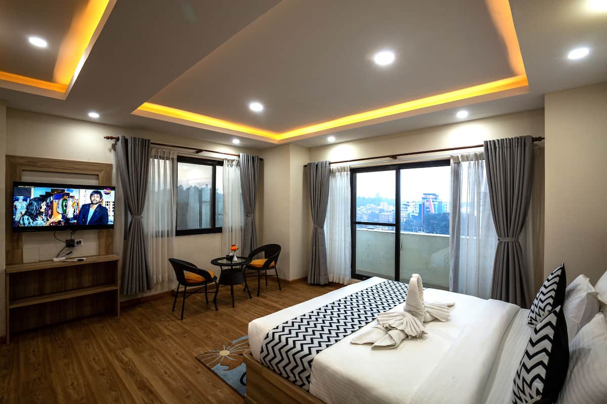 A hotel room with a large bed, a TV, a desk with two chairs, and a view of the city through the windows.