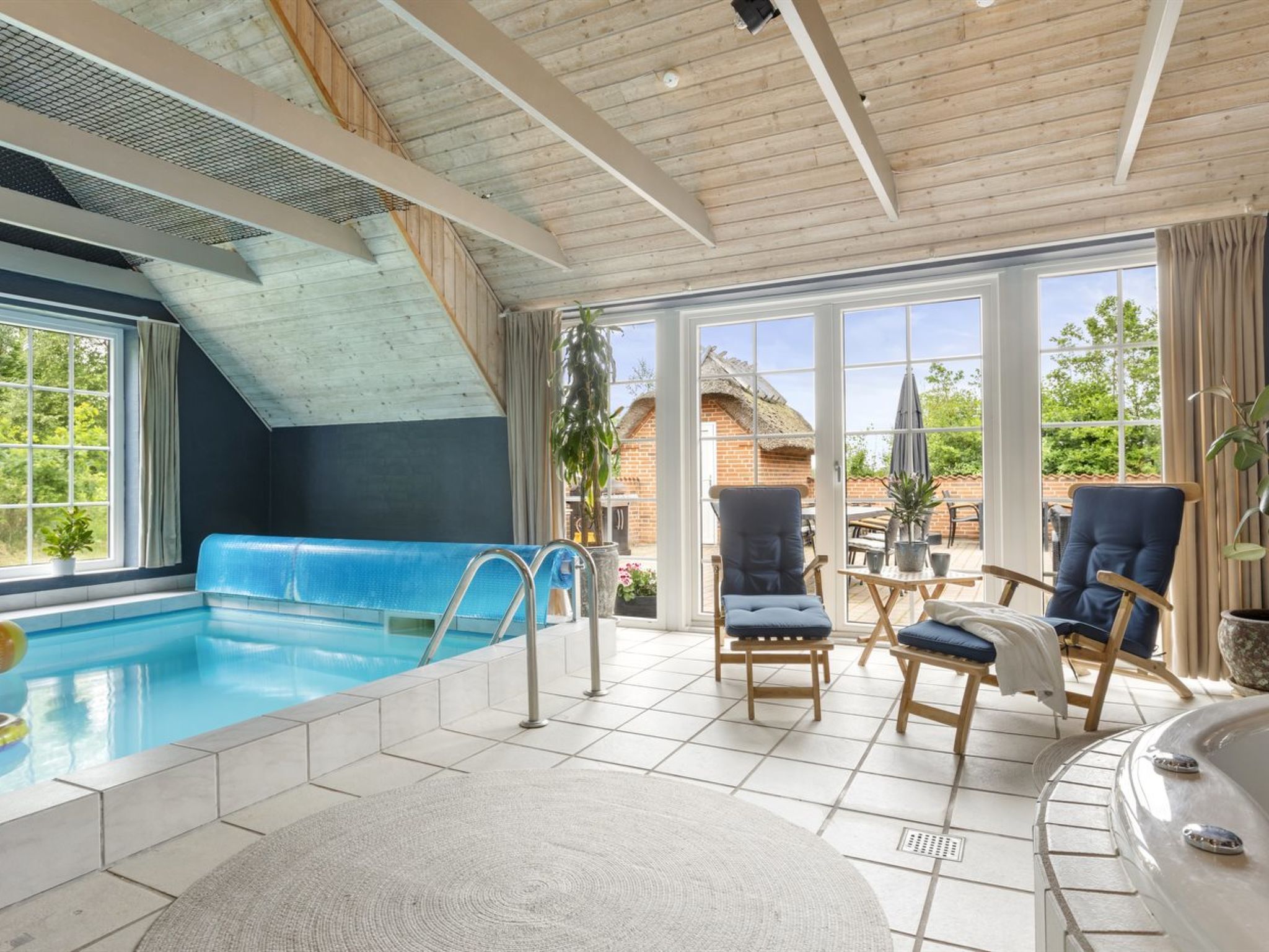 Ett poolområde med en blå pool, solstolar och utsikt över ett hus genom stora fönster.