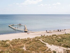 A beach with a wooden pier extending into the water, surrounded by sand and grassy dunes.