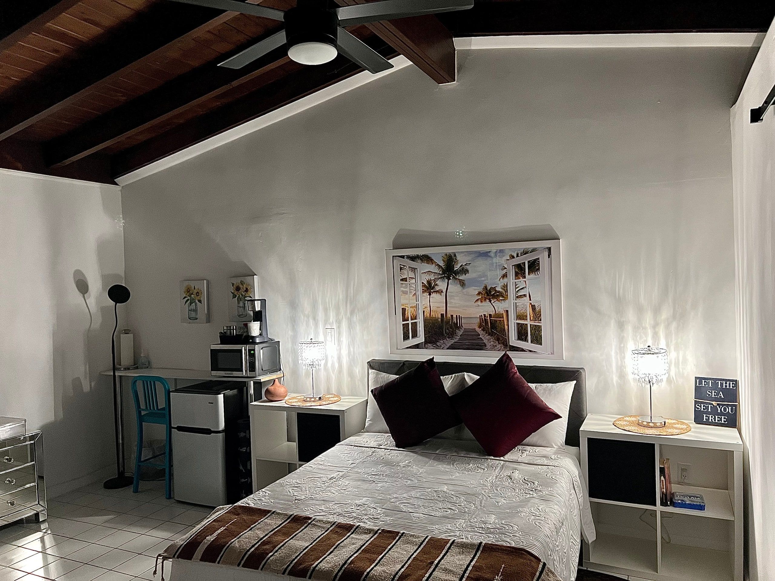 A bedroom with a bed, a desk, a chair, a microwave, and a nightstand with a lamp.