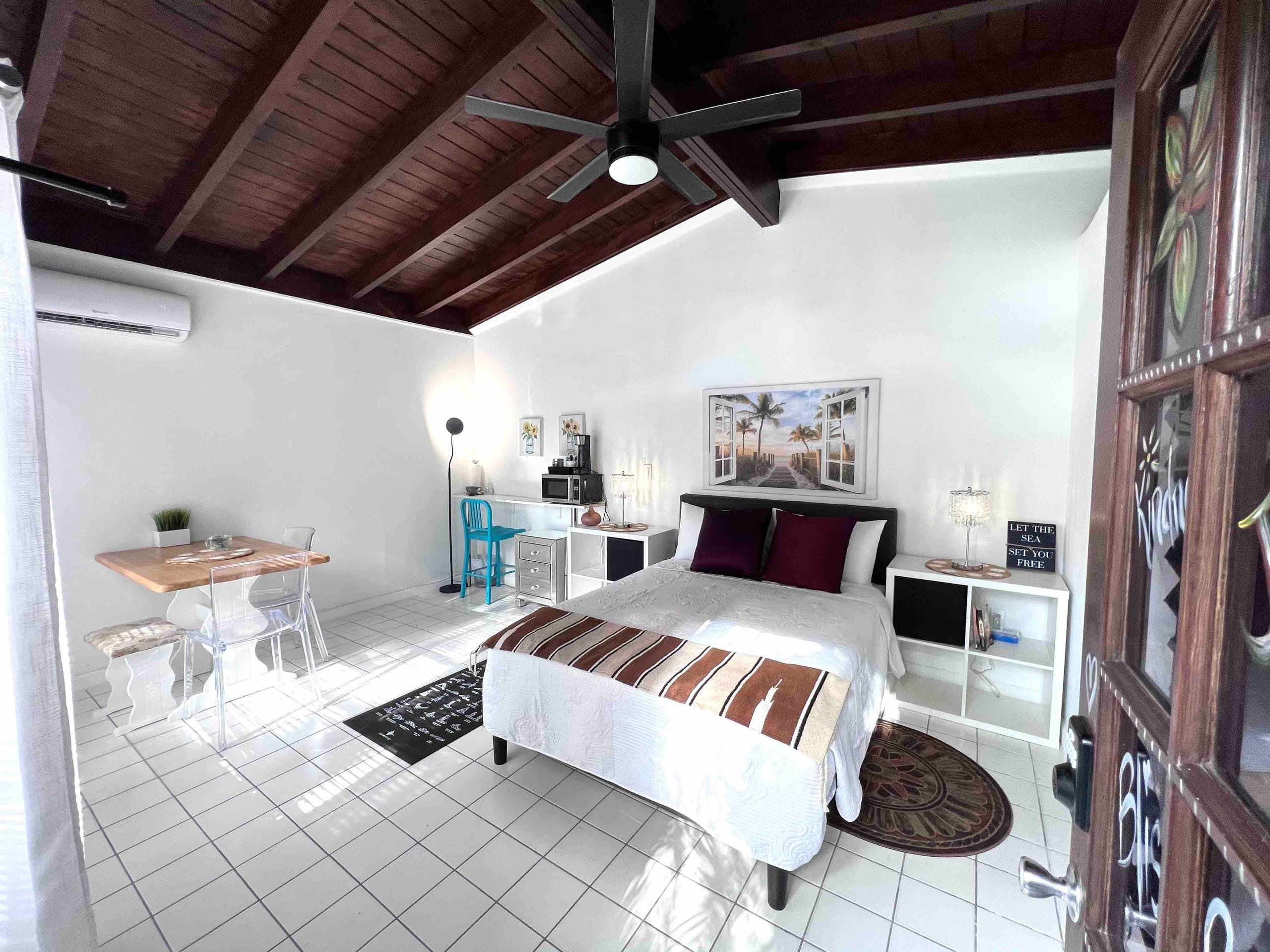 Un dormitorio con cama, escritorio, silla, mesita de noche, ventilador de techo y ventana.