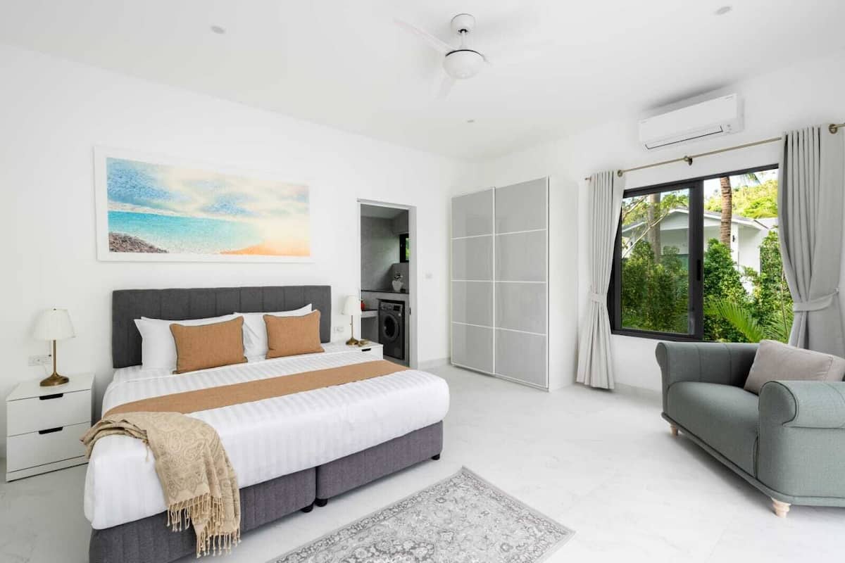 Un dormitorio moderno con una cama grande, un sofá gris, vistas a zonas verdes y un cuadro en la pared.