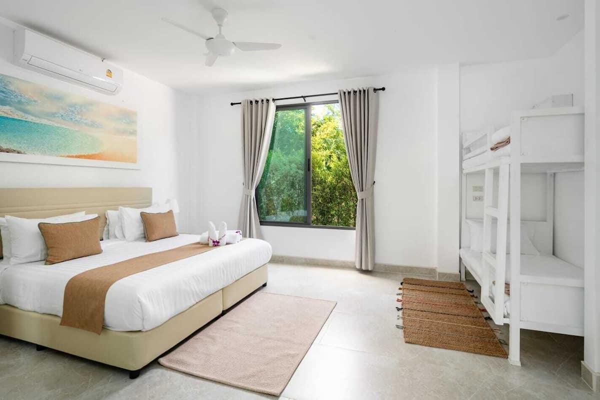 Habitación de hotel con cama, ventilador de techo, ventana con cortinas, un cuadro en la pared y una litera a la derecha.