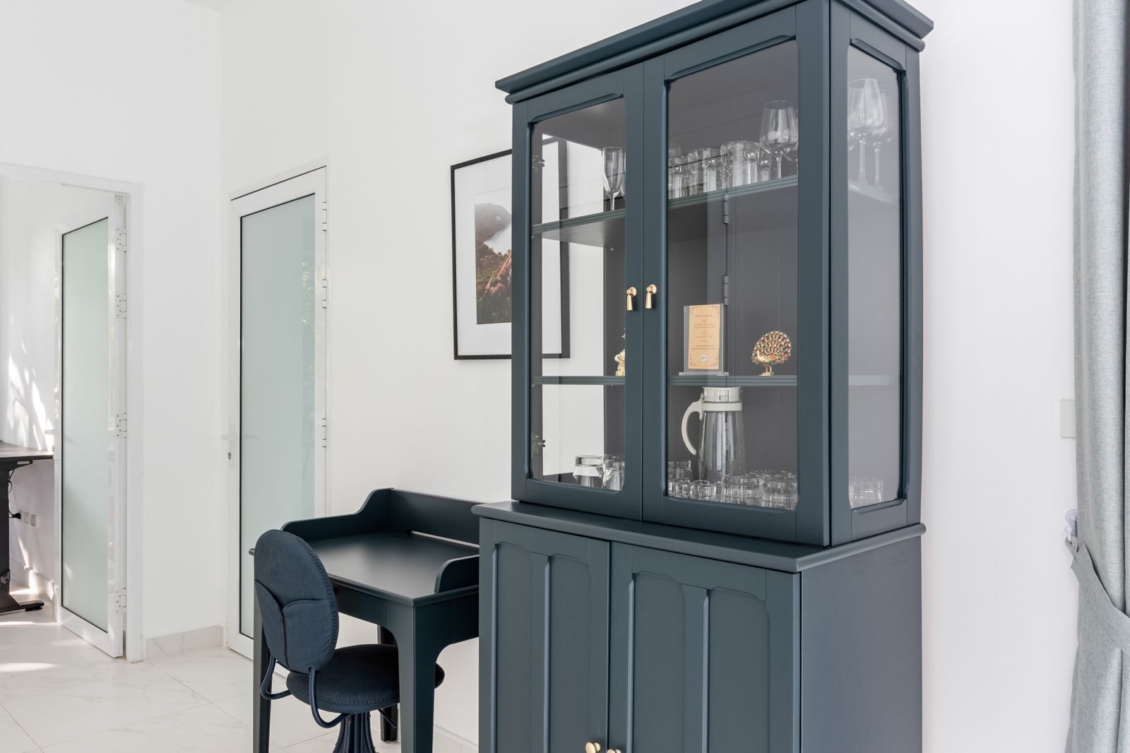 A room with a dark blue cabinet containing glassware and a coffee maker, a black chair, and a framed picture on the wall.