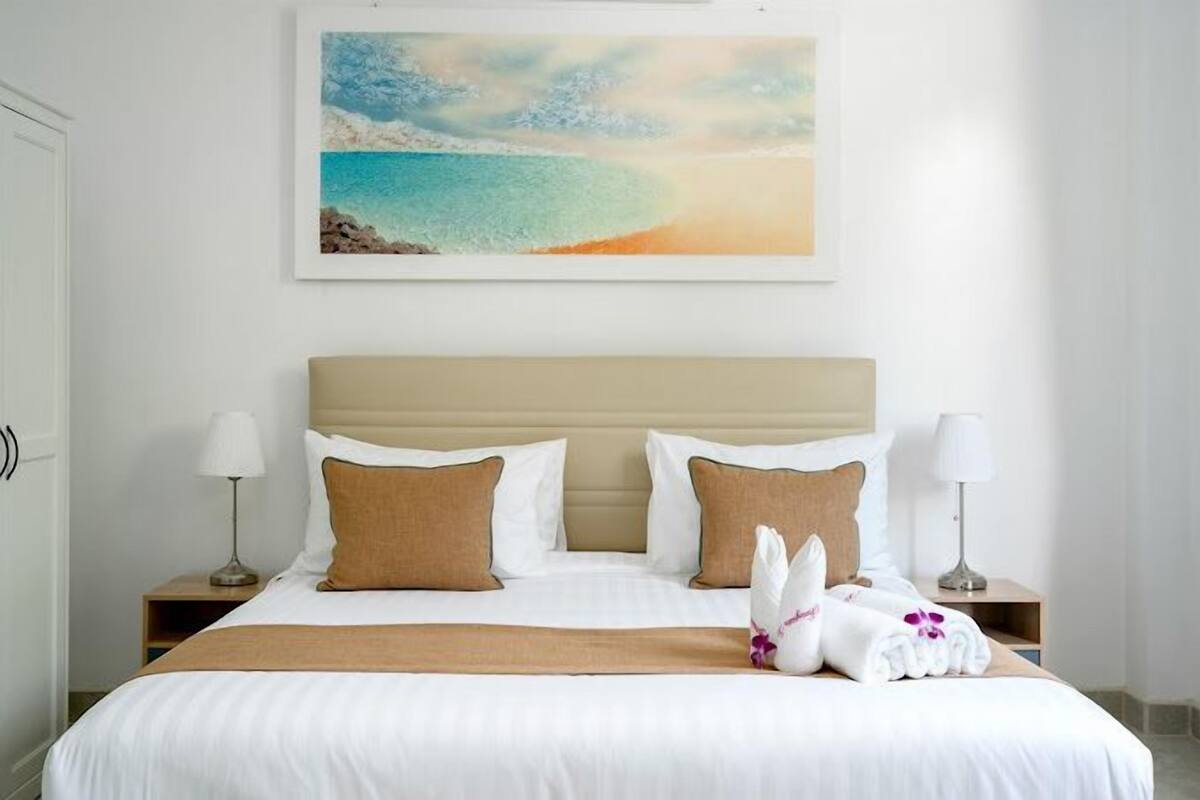 Habitación de hotel con cama, dos mesitas de noche, cabecera y un cuadro de una playa.