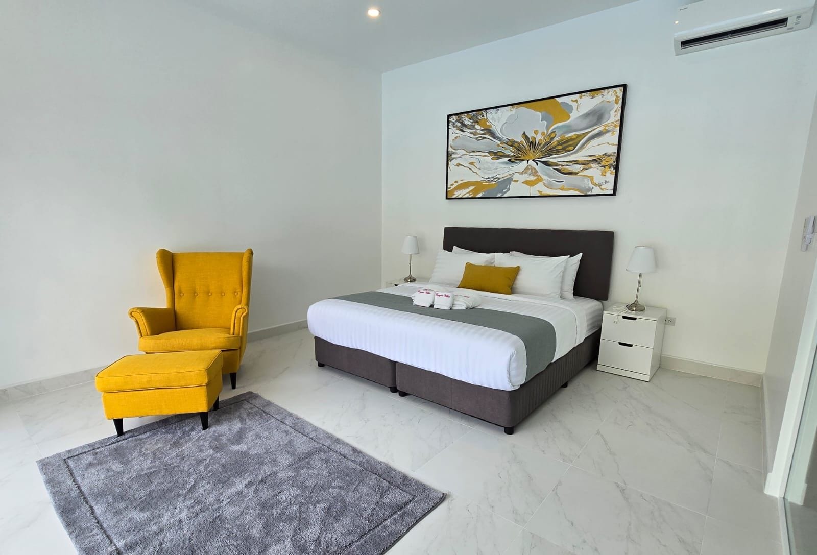 Un dormitorio moderno con una cama, un sillón amarillo, una alfombra gris y un cuadro en la pared.