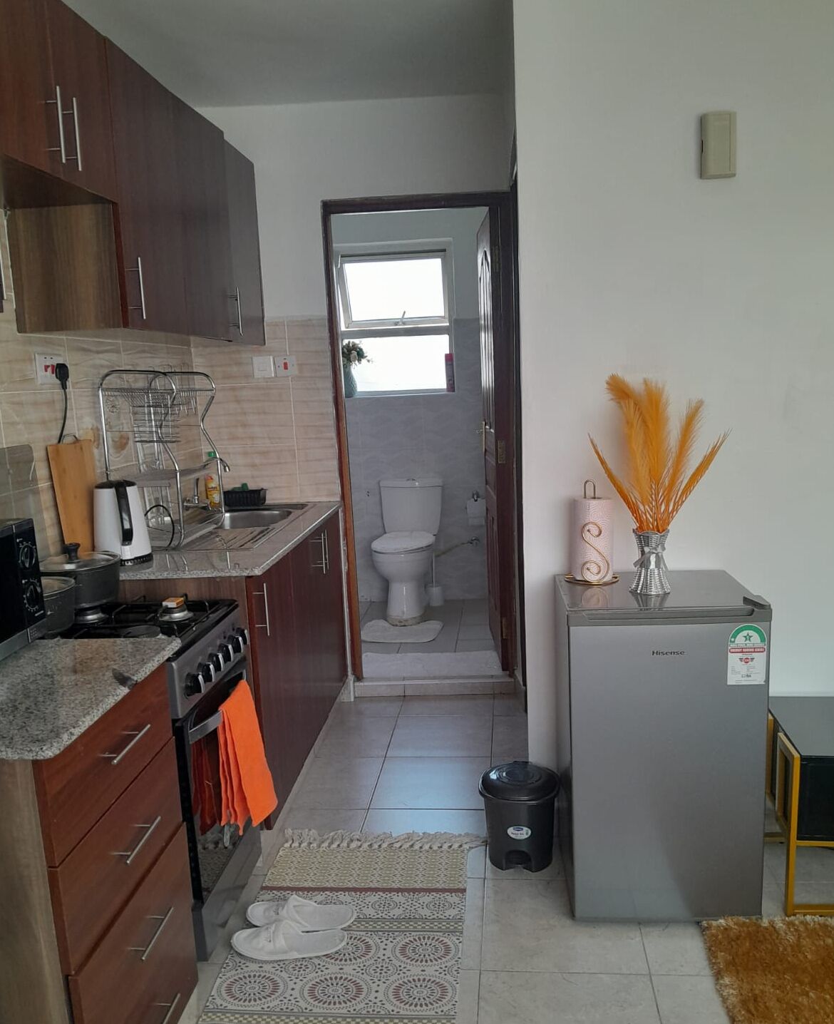 Cocina compacta con estufa, horno y lavabo, que da paso a un baño pequeño con inodoro y ventana.