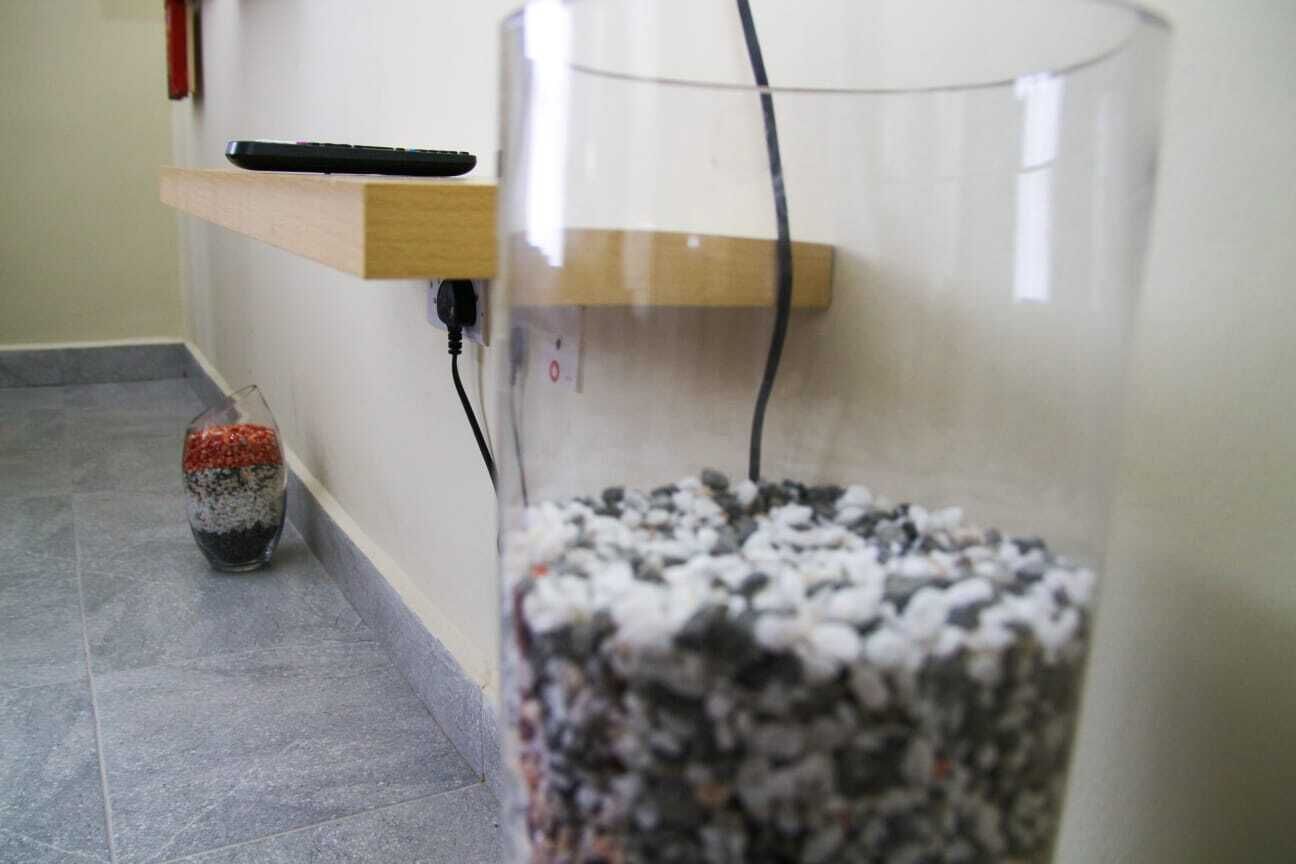 A room with a glass aquarium on the floor, a wooden shelf with a remote control, and a power outlet on the wall.