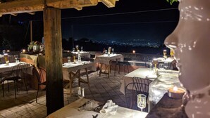 An outdoor dining area with tables set for two, under a wooden pergola, overlooking a cityscape at night.