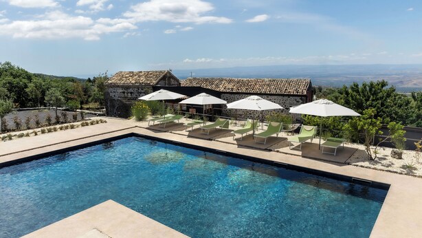 A swimming pool with lounge chairs and umbrellas, surrounded by a stone building with a tiled roof, overlooking a scenic landscape.