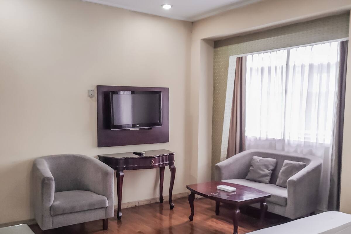 A hotel room with a bed, a sofa, a chair, a small table, a TV mounted on the wall, and a window with curtains.