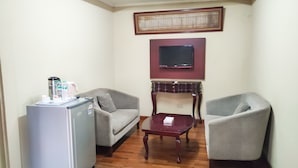 Uma sala compacta com geladeira, dois poltronas, uma mesa de madeira pequena e uma televisão fixada na parede.