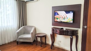 Uma sala de estar com uma poltrona cinza, uma mesa redonda pequena, uma televisão fixada na parede e um aparelho de ar condicionado.