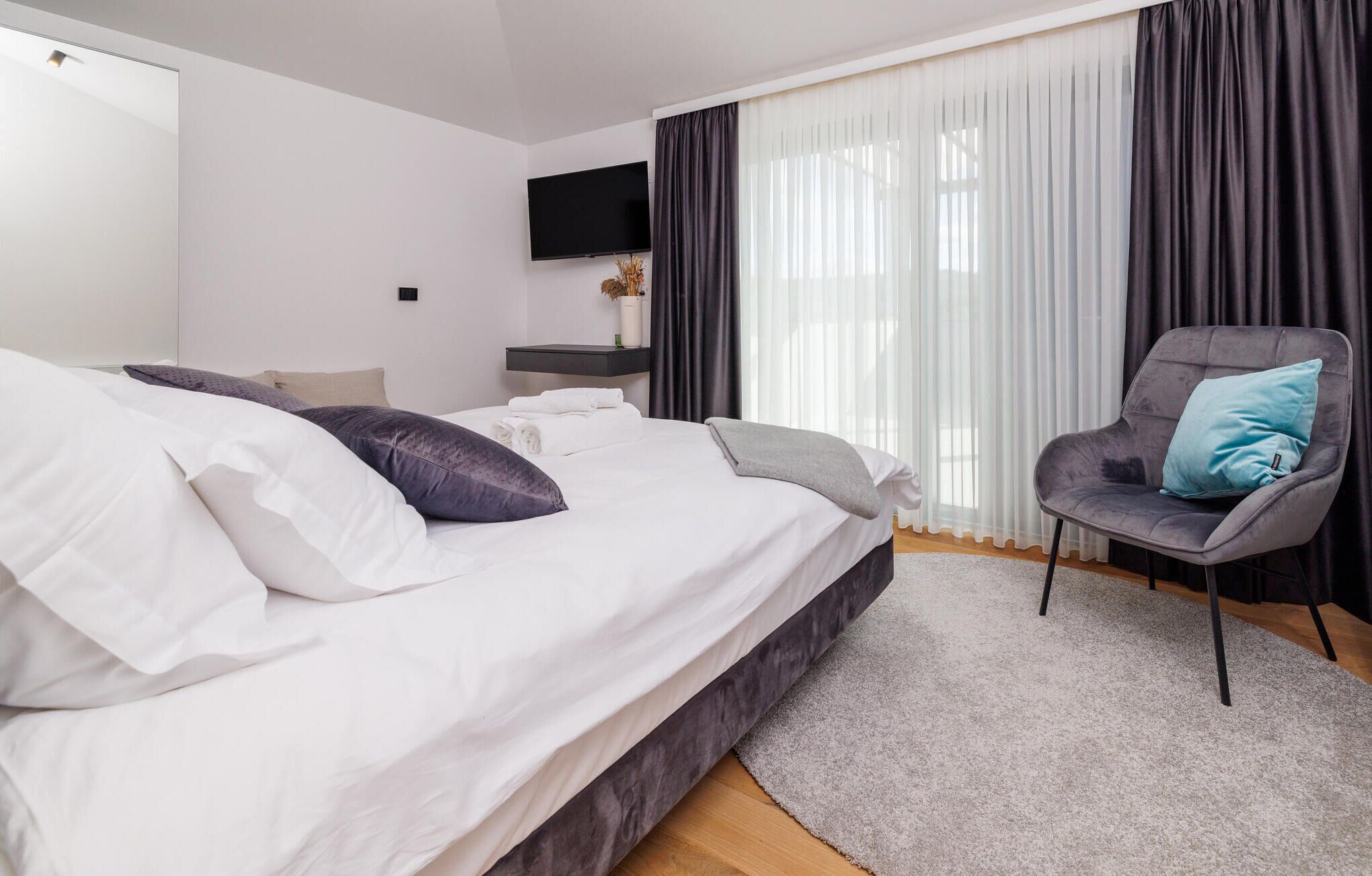 Une chambre moderne avec un grand lit, un fauteuil gris, un téléviseur à écran plat et une fenêtre ornée de rideaux légers.