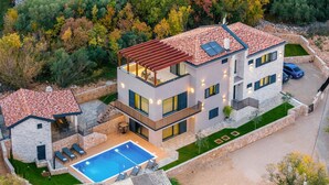 A modern two-story house with a swimming pool, stone walls, and a view of a forest.