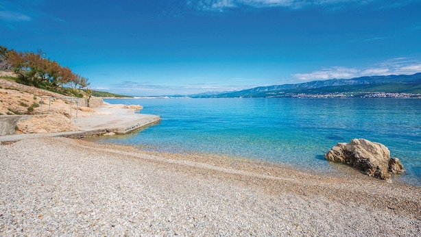 A pebbled beach with clear blue water, a concrete walkway, and rocky terrain with sparse vegetation.